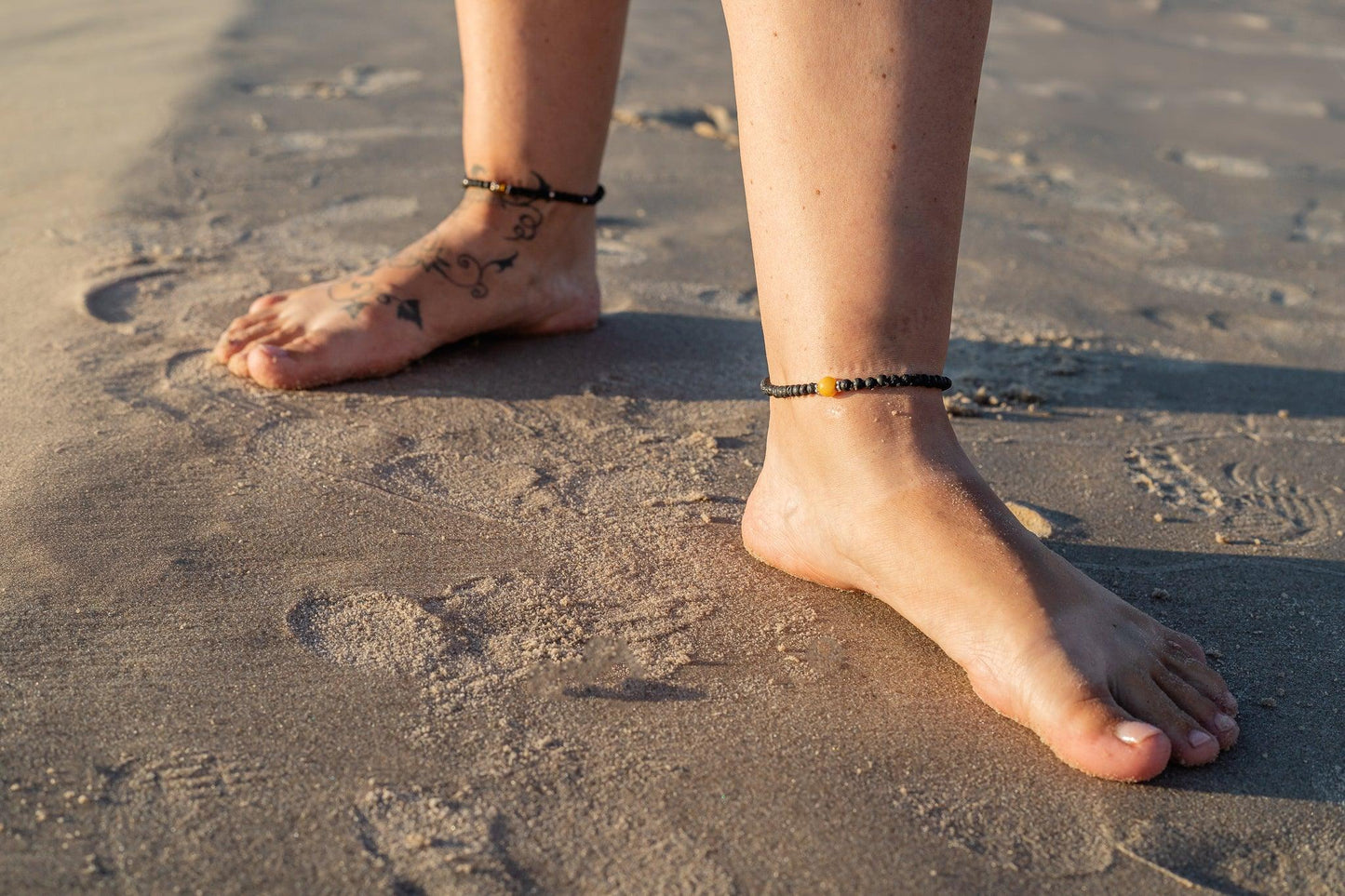 Lava stone anklet bracelet with green Baltic amber gem on sandy beach.