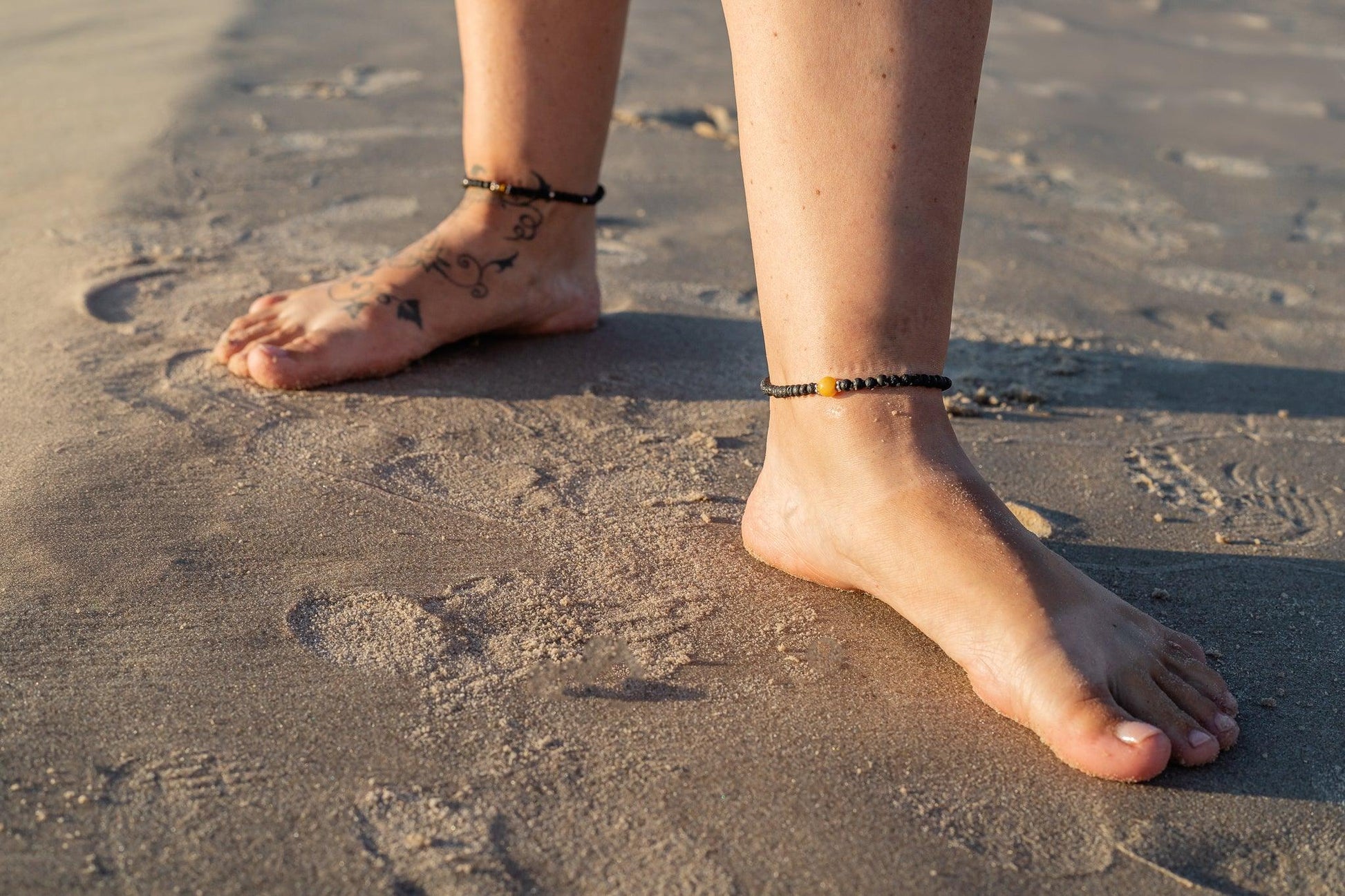 Lava stone anklet bracelet with green Baltic amber gem on sandy beach.