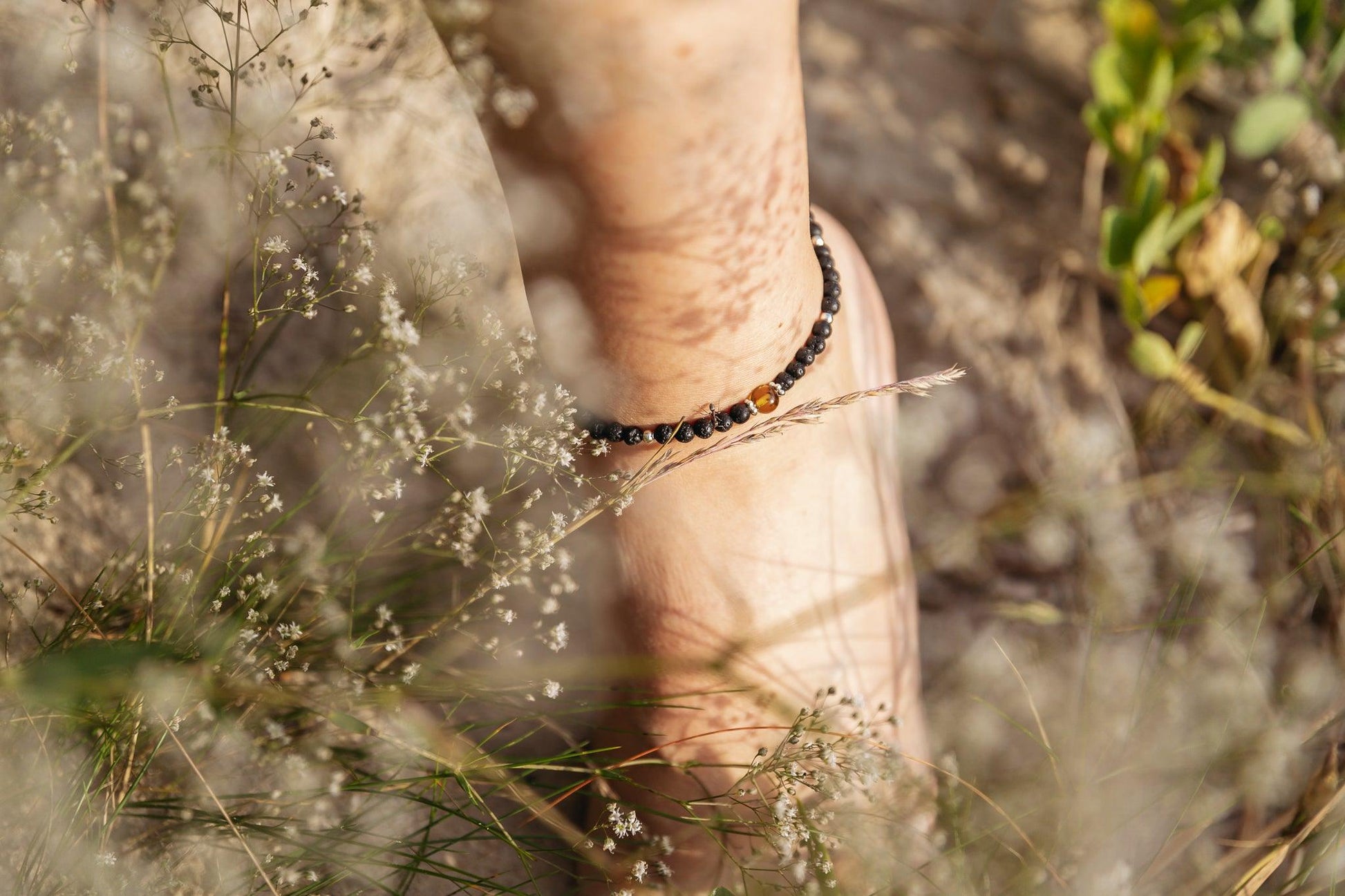 Lava stone anklet bracelet with green Baltic amber gem on ankle surrounded by grass and flowers.